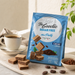 A bag of Lago Be Goodie Sugar Free Mini Party Cacao Cocoa wafer cookies stands on a light stone surface beside a cup of coffee, scattered wafer pieces, cocoa powder, and a small bowl, with a soft neutral background and plant leaves at the edge.