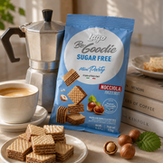 A blue bag of Lego Be Goodie Sugar Free Mini Party hazelnut cookies stands on a tabletop beside a white coffee cup, stacked books, plates of cookies, and a silver moka pot. The bag shows the Lego logo and product text, with cookies and hazelnuts pictured on the packaging.