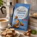 A blue bag of Lego Be Goodie Sugar Free Mini Party hazelnut cookies stands on a tabletop beside a white coffee cup, stacked books, plates of cookies, and a silver moka pot. The bag shows the Lego logo and product text, with cookies and hazelnuts pictured on the packaging.