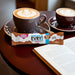 Two cups of coffee with a bar of EVRY1 sugar-free milk chocolate on a table next to an open book.