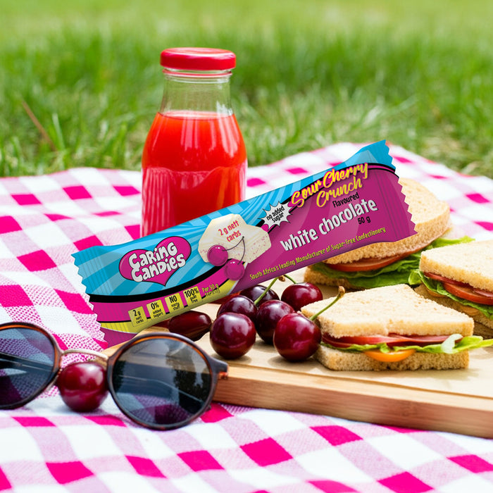 Picnic setup with sugar-free caring candies white chocolate bar, juice, sandwiches, and cherries on a checkered blanket.