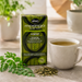 A box of Akan Moringa Leaf Tea stands on a stone surface beside a cup of brewed tea and scattered loose leaves, with blurred plants in the background.