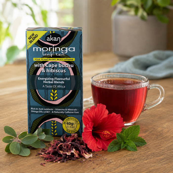 A box of Akan Moringa Leaf Tea is displayed on a wooden table next to a glass of brewed dark red tea, dried hibiscus flowers, fresh green leaves, and a red hibiscus flower. The background is a softly blurred kitchen setting.