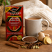 A box of Akan Moringa Leaf Tea with Cape Foöbois Chai is displayed on a wooden table alongside a cup of brewed tea, ginger root, cinnamon sticks, star anise, and cardamom pods, with a cozy knitted blanket in the background.