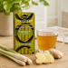 A box of Akan Moringa Leaf Tea stands on a wooden table beside a clear glass mug filled with the tea, along with lemongrass stalks, ginger slices, and ginger root. The background is softly blurred with green leaves and a neutral indoor setting.