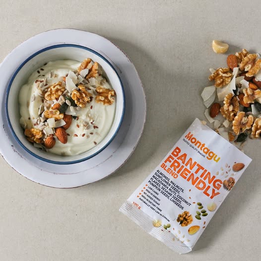 A bowl of white yogurt topped with walnuts, almonds, and coconut flakes sits on a white plate beside a packet of Montagu Banting Friendly snack blend, with some of the snack mix spilled on a light beige surface.