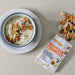 A bowl of white yogurt topped with walnuts, almonds, and coconut flakes sits on a white plate beside a packet of Montagu Banting Friendly snack blend, with some of the snack mix spilled on a light beige surface.
