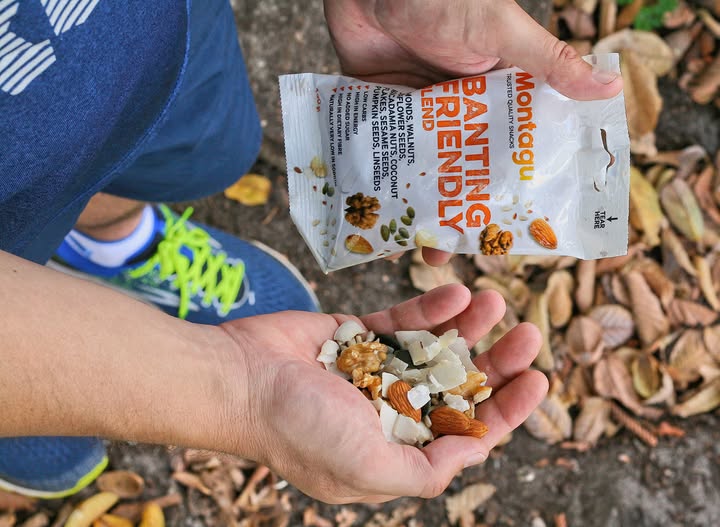 A person outdoors holds an open bag of Monotugu Banting Friendly snack mix over their open palm, showing the nuts and coconut pieces. The background shows a natural ground with dry leaves.