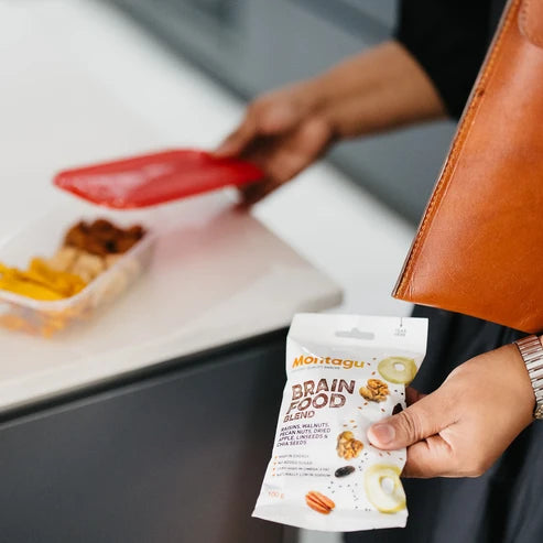 A person holds a white bag of Maitagu Brain Food Blend in one hand while carrying a brown leather folder in the other. In the blurred background, a hand uses a red spatula to mix food in a container on a white counter.