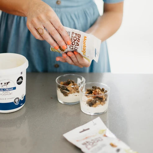 Person adding toppings to yogurt with a container of granola and a carton of yogurt on a table.