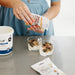 Person adding toppings to yogurt with a container of granola and a carton of yogurt on a table.