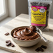 A bowl of chocolate compound milk chocolate spread sits on a stone surface with a spoon, alongside a clear stand-up pouch of Caring Candies compound milk chocolate chips. The pouch has a yellow and pink label, and several chocolate pieces are scattered around.