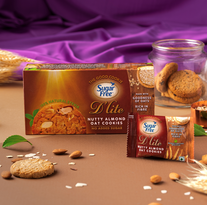 Sugar Free D-lite Nutty Almond Oat Cookies packaging shown with a large box and a single packet on a brown surface, with cookies and a glass jar of cookies in the background and purple fabric behind them.