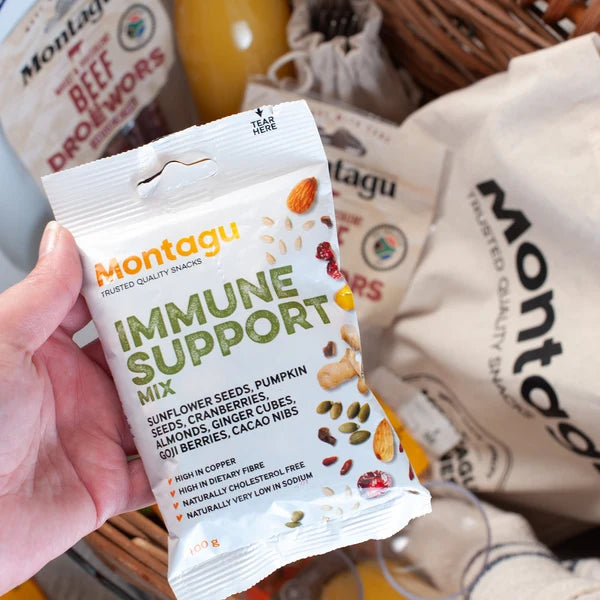 A hand holds a bag of Montagu Immune Support Mix in front of other Montagu snack bags and a wicker basket, showing the packaged snack product in a real setting.