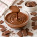 A glass bowl filled with smooth chocolate, with a spoon lifting some of the chocolate above the bowl. Broken chocolate pieces are scattered around on a light marble surface, and a small white ramekin of chocolate chunks sits in the background.