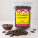 A clear stand-up pouch of Caring Candies No Added Sugar Compound Milk Choc chocolate pieces stands on a white marble surface with a small bowl of milk-chocolate sauce and scattered chocolate chunks in front. The front label is bright yellow and pink with product text, and the pouch is photographed against a clean white studio background.