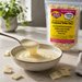 A bag of Caring Candies No Added Sugar Compound White Choc in a kitchen-like setting with a bowl of melted white chocolate and a spoon dripping chocolate, with scattered white chocolate pieces around.