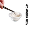 A spoonful of xanthan gumpowdery substance, gluten-free flour, is being measured with a spoon on a clear glass bowl. One hand is holding the spoon, and the other is adjusting the measure on the spoon against the bowl.