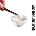 A spoonful of xanthan gumpowdery substance, gluten-free flour, is being measured with a spoon on a clear glass bowl. One hand is holding the spoon, and the other is adjusting the measure on the spoon against the bowl.