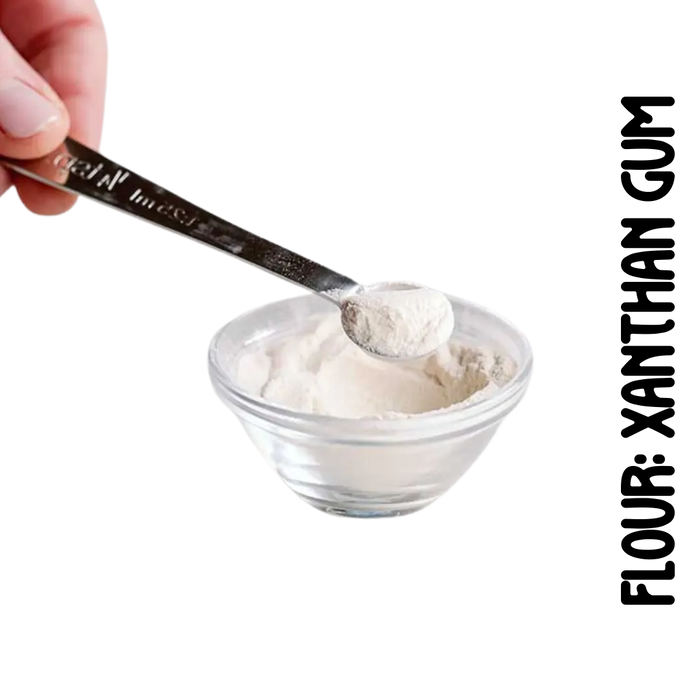 A spoonful of xanthan gumpowdery substance, gluten-free flour, is being measured with a spoon on a clear glass bowl. One hand is holding the spoon, and the other is adjusting the measure on the spoon against the bowl.