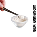 A spoonful of xanthan gumpowdery substance, gluten-free flour, is being measured with a spoon on a clear glass bowl. One hand is holding the spoon, and the other is adjusting the measure on the spoon against the bowl.
